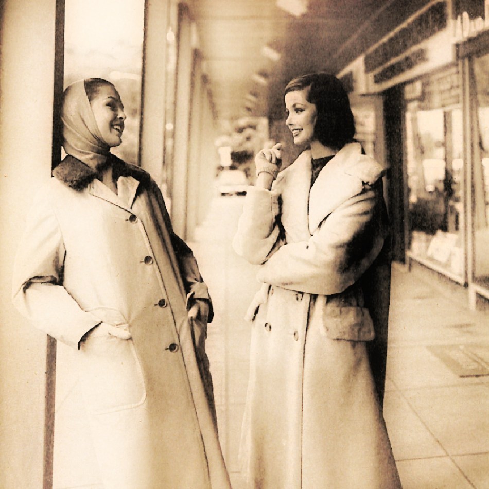 2 1950s women wearing coats talk in the front of a shopping center.